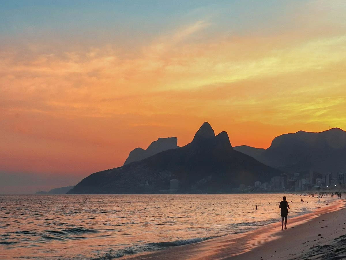 A retired couple walking along a beach in Brazil during a golden sunset with mountains in the background.