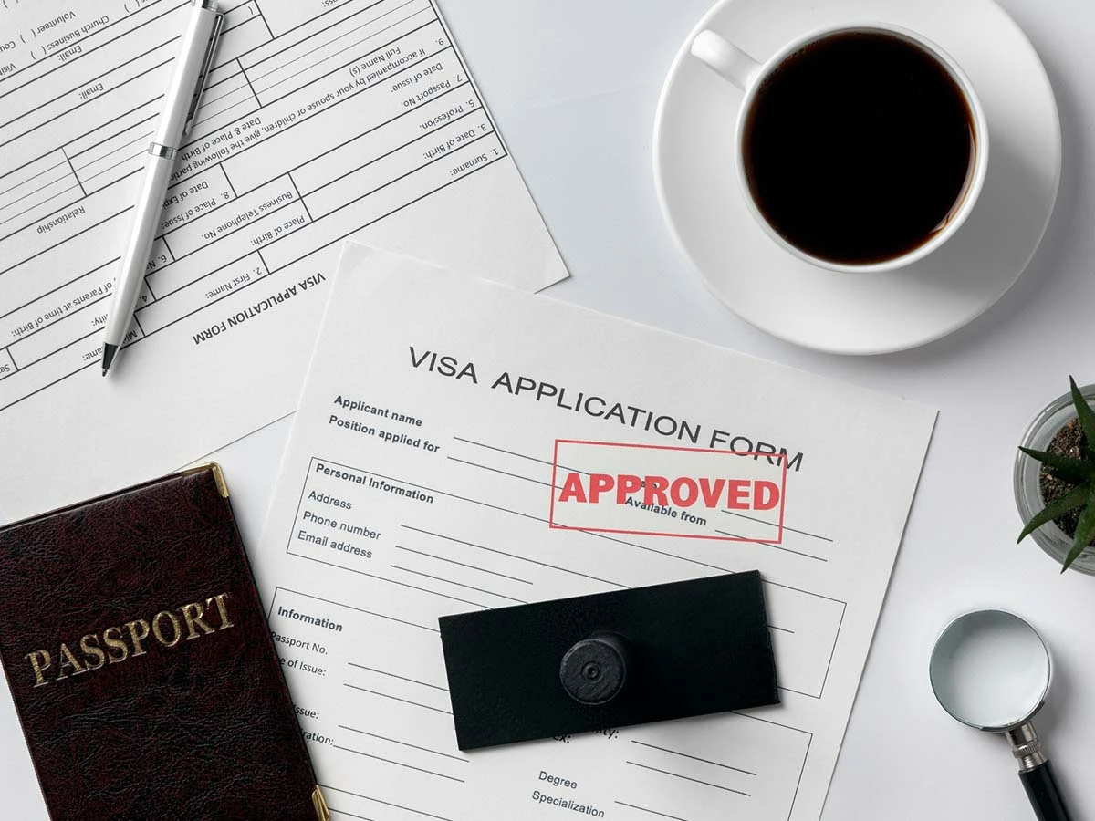A top-down view of a visa application form with a red "Approved" stamp next to a passport and coffee.