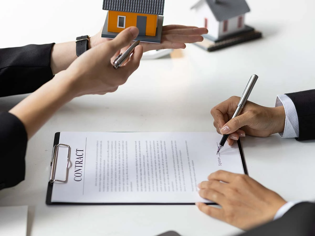 Close-up of a hand signing a legal real estate contract next to a small house model, illustrating the property buying process in Brazil.