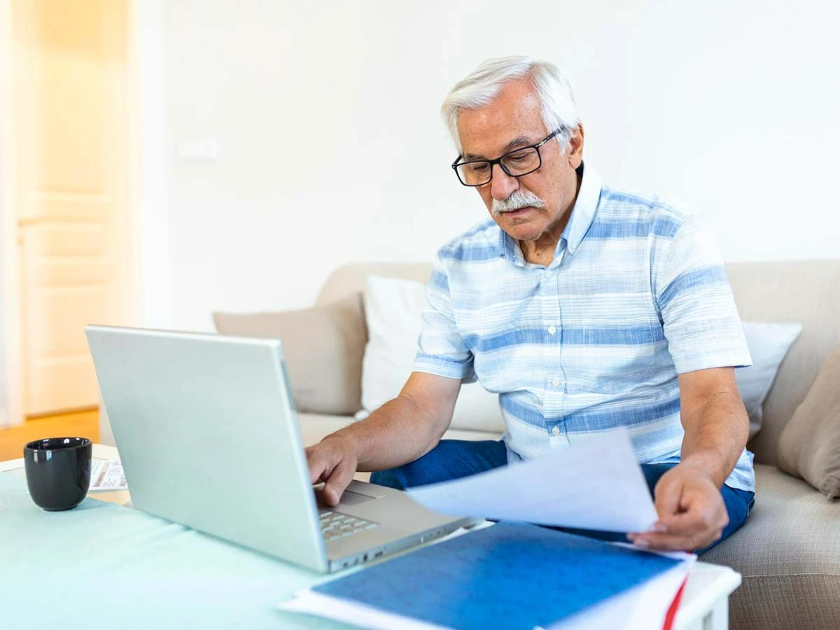 A senior man sitting on a sofa using a laptop and reviewing financial bank statements for his retirement budget.